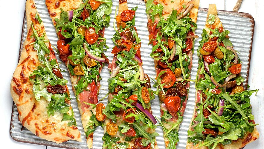 Eine große flache Pizza mit knusprigem Rand, belegt mit frischem Rucola, getrockneten Tomaten und roten Zwiebeln. Die Pizza ist in gleichmäßige Stücke geschnitten und auf einem gestreiften Blech angerichtet, was einen appetitlichen Anblick bietet.