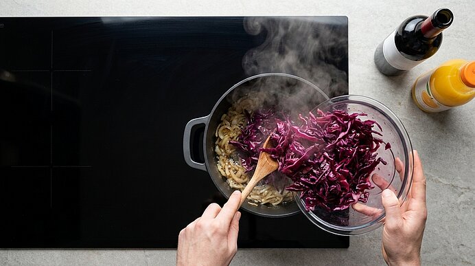 Hände geben geschnittenen Rotkohl aus einer Glasschüssel in einen dampfenden Kochtopf; ein Holzlöffel rührt angebratene Zwiebeln auf dem Herd.