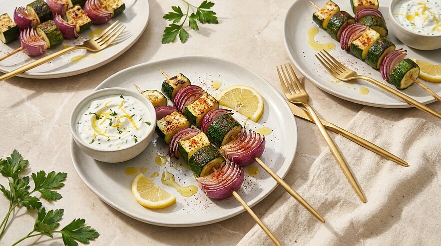 Zwei Grillspieße mit Halloumi, Zucchini und roten Zwiebeln auf einem Teller, serviert mit Zitronenscheiben und einer Schale Joghurt-Dip, garniert mit Petersilie.
