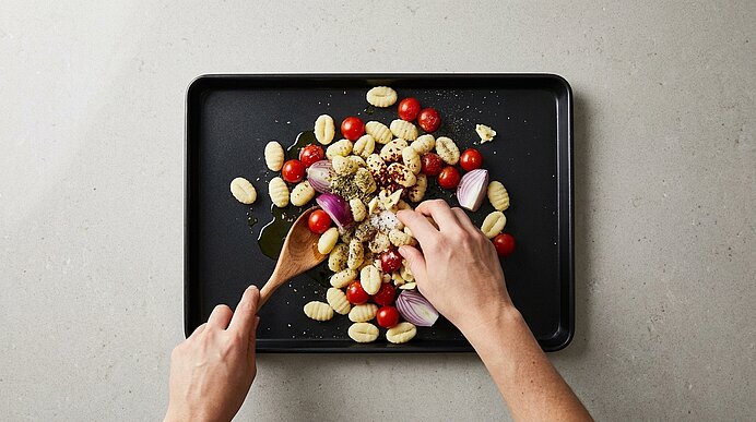 Zwei Hände vermengen Gnocchi, Kirschtomaten und rote Zwiebelhälften auf einem Backblech, beträufelt mit Olivenöl und Kräutern.