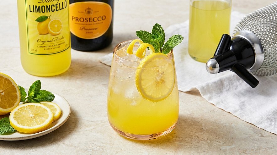 Glas mit gelbem Limoncello‑Prosecco‑Cocktail, Eis, Zitronenscheibe, Zitronenzeste und Minze; daneben Limoncello‑ und Prosecco‑Flaschen sowie Zitronenscheiben.