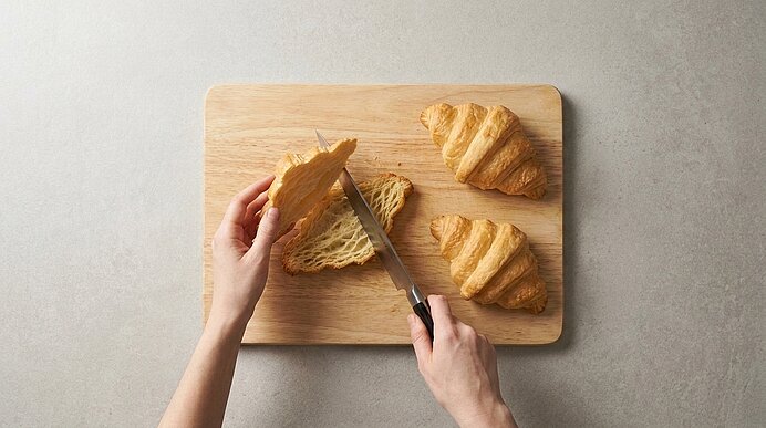 Zwei Hände schneiden ein Croissant auf einem Holzbrett waagerecht durch. Daneben liegen zwei weitere Croissants.