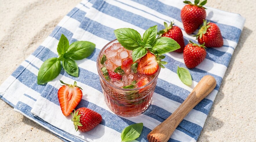 Glas mit eisgekühltem Erdbeer-Basilikum-Getränk, garniert mit Basilikumblatt und Erdbeeren, umgeben von frischen Erdbeeren und Basilikumblättern auf blau-weiß gestreiftem Tuch.