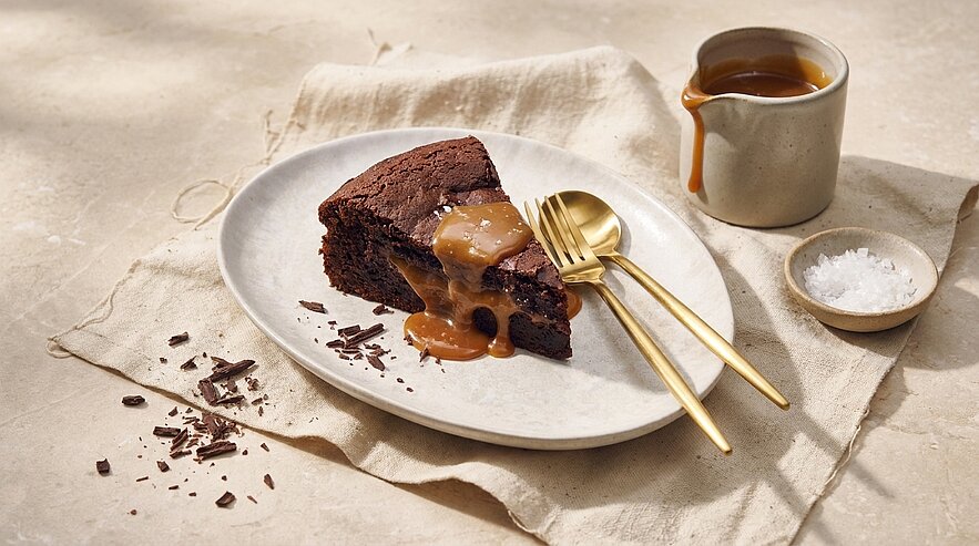 Ein saftiges Stück Schokoladenkuchen auf ovalem Teller, mit zähflüssiger Karamellsauce übergossen; daneben goldene Gabel und Löffel, kleine Kanne mit Karamell, Schale Meersalz und Schokoladenraspel.