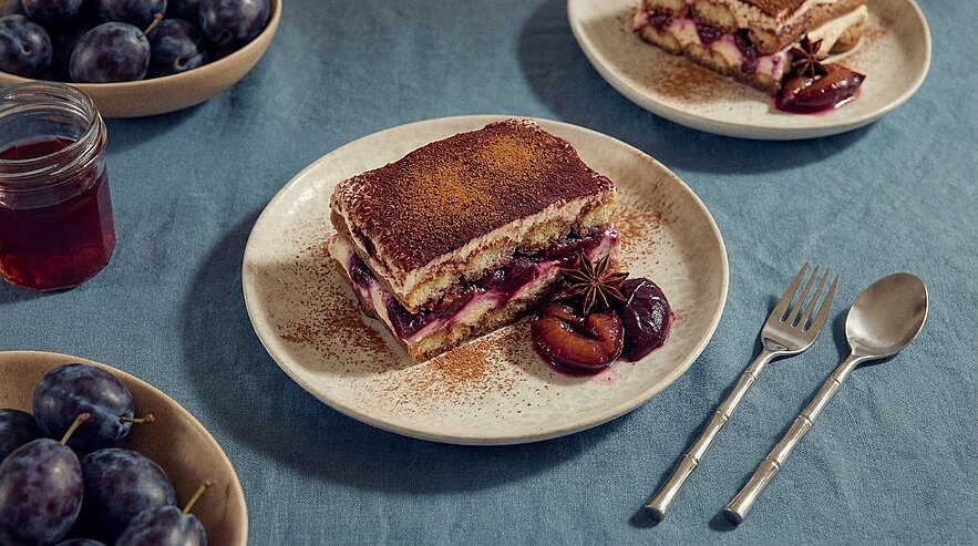 Scheibe Schichtdessert mit Pflaumenfüllung und Kakaopulver auf einem Teller, garniert mit halben Pflaumen und Sternanis, auf blauem Tischtuch.