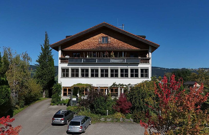 Ein mehrstöckiges Haus mit einem Satteldach, umgeben von Bäumen und buntem Laub. Im Vordergrund sind zwei parkende Autos auf einem gepflasterten Weg zu sehen. Der Himmel ist klar und blau, was auf einen sonnigen Tag hinweist.