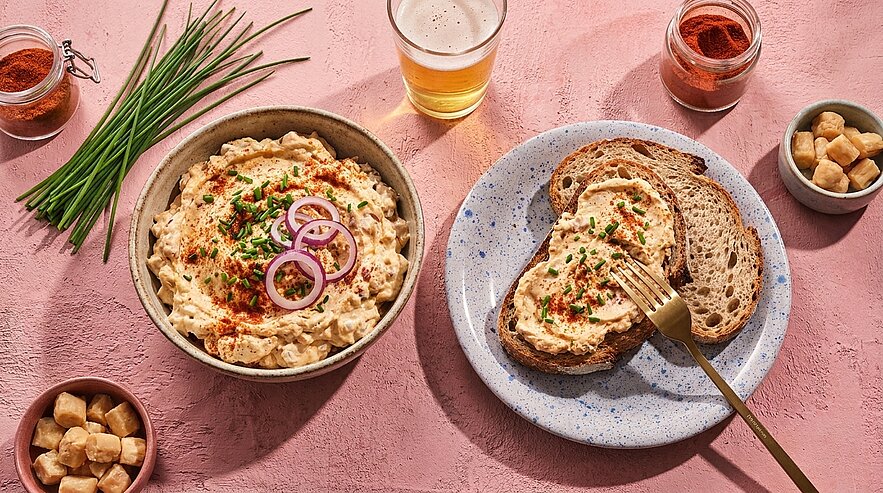 Schale mit cremigem Käseaufstrich, garniert mit Schnittlauch und roten Zwiebelringen; auf dem Teller zwei Scheiben Vollkornbrot, eine mit dem Aufstrich bestrichen und Gabel.