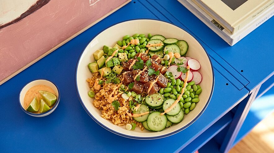 Schüssel mit farbenfroher Poké-Bowl: gewürfelter roher Fisch, Gurkenscheiben, Radieschen, Edamame, Avocadowürfel, Körner (Bulgur), Frühlingszwiebeln und Sesam; daneben kleine Schale mit Limetten und Dip auf blauem Tisch.