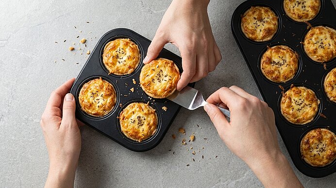 Eine goldbraun gebackene Mini-Chäswähe wird mit einem kleinen Heber aus der dunklen Muffinform gehoben.