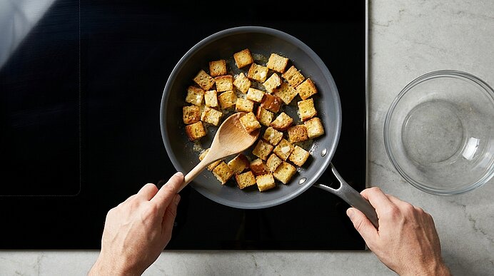 Zwei Hände rühren mit einem Holzlöffel goldbraune Brotwürfel (Croutons) in einer Pfanne auf dem Herd.