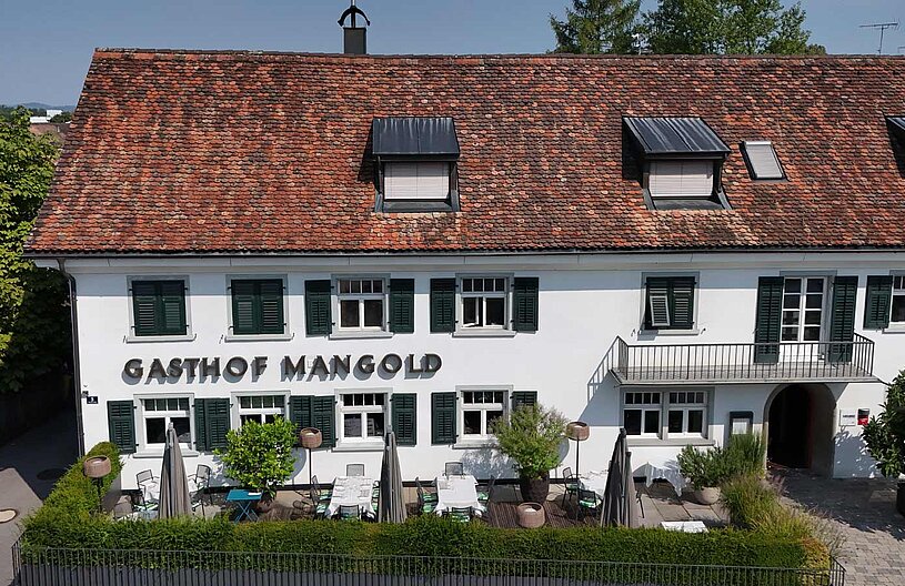 Ein traditionelles Gasthaus mit weißer Fassade und einem roten Ziegeldach. Der Name "Gasthof Mangold" ist sichtbar angebracht. Vor dem Gebäude befinden sich Gartentische, Stühle und Sonnenschirme, umgeben von grünen Pflanzen.