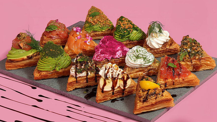Eine Teller mit mehreren dreieckigen Blätterteigtörtchen, gefüllt mit verschiedenen Zutaten wie Lachs, Avocado, rosa Creme und Sahne. Einige sind mit Kräutern und Nüssen garniert. Der Hintergrund ist rosa, und die Törtchen sind auf einer dunklen Platte angeordnet.
