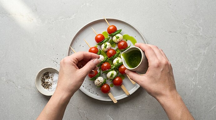 Hände beträufeln Caprese-Spieße auf einem Teller mit Basilikumöl und würzen sie mit Salz und Pfeffer.