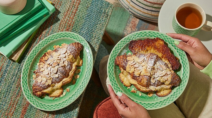 Zwei goldbraune Mandelcroissants auf grünen Tellern, eines wird von Händen gehalten; mit Mandelblättchen und Puderzucker bestreut, daneben eine Tasse Tee.