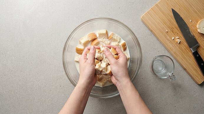 Zwei Hände drücken eingeweichte Brötchenwürfel über einer Glasschüssel mit Milch aus.