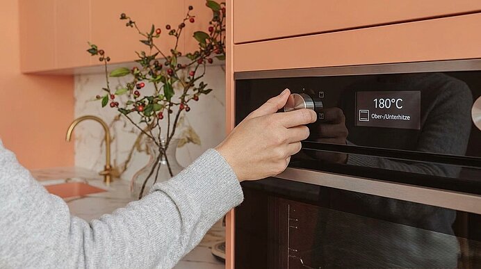 Hand einer Person in grauem Pullover dreht den Temperaturregler an einem eingebauten Ofen; digitales Display zeigt 180 °C in einer modernen Küche.