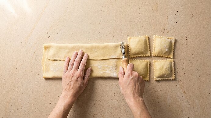 Hände schneiden mit einem Teigrädchen eine Teigbahn zu quadratischen Ravioli; rechts liegen vier fertig geformte, gefüllte Ravioli auf der Arbeitsfläche.