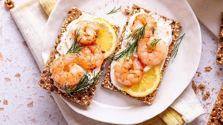 Ein Teller mit zwei knusprigen Brotstücken, belegt mit Sahne, garnierte Garnelen, Zitronenscheiben und frischem Dill. Der Teller befindet sich auf einer hellen Oberfläche, daneben liegt ein saftiges Tuch.