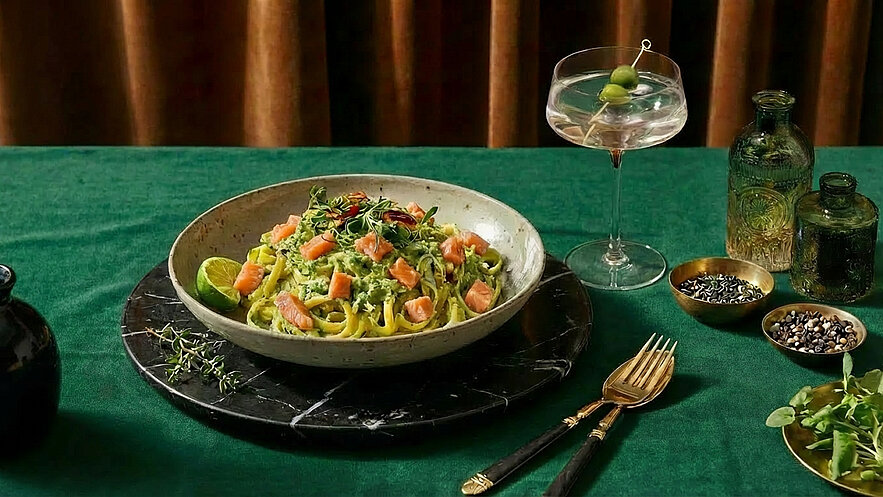 Ein Teller mit frischer Pasta, garniert mit Stücken von Lachs und Kräutern, steht auf einem schwarzen Marmorteller. Daneben befindet sich ein Glas Martini mit Oliven. Der Tisch ist mit einem grünen Tuch bedeckt.