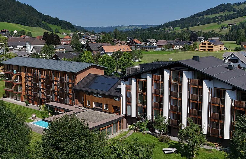 Ein modernes, mehrstöckiges Gebäude mit großen Fenstern und Balkonen steht in einer grünen Landschaft. Im Vordergrund befindet sich ein Schwimmbereich, umgeben von gepflegtem Gras und Bäumen. Im Hintergrund sind einige traditionelle Häuser und sanfte Hügel sichtbar.