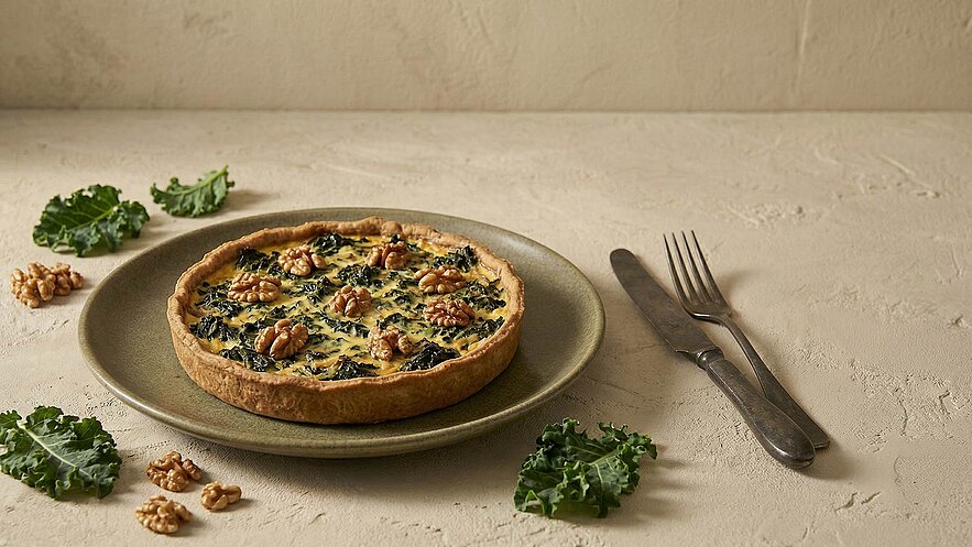Eine herzhafte Quiche mit einem Mürbeteigboden, gefüllt mit grünem Blattgemüse und Walnüssen. Sie liegt auf einem flachen Teller. Einige frische Grünkohlblätter und Walnüsse sind um den Teller angeordnet, und Besteck liegt daneben.