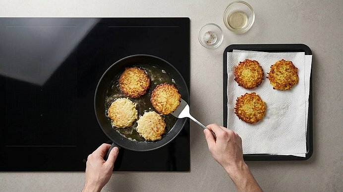 Zwei Hände wenden Kartoffelpuffer in einer Pfanne. Rechts liegen fertige goldbraune Puffer auf Küchenpapier.