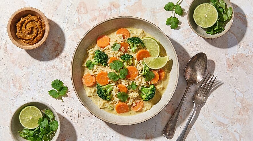 Schale mit cremigem Gemüse-Curry und Reis, garniert mit Brokkoli, Karottenscheiben, Koriander und Limettenspalten; Löffel und Gabel daneben.