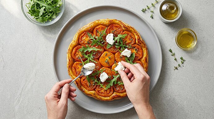 Gebackene Tomatentarte auf einem Teller; Hände setzen kleine Käsetupfen und Rucola als Garnitur.