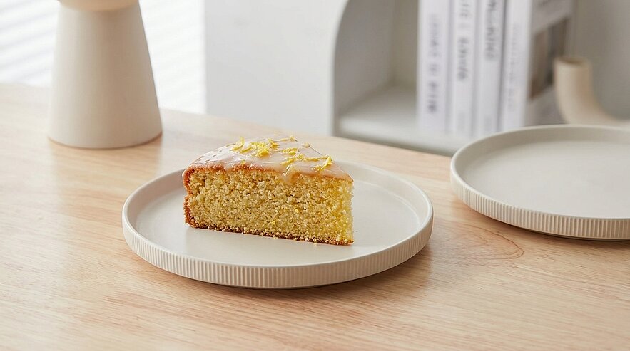Ein Stück Zitronenkuchen auf einem hellen, runden Teller, platziert auf einem Holzgestell. Der Kuchen hat eine glasiierte Oberfläche und ist mit Zitronenschalen dekoriert. Im Hintergrund sind unscharfe Bücher sichtbar.