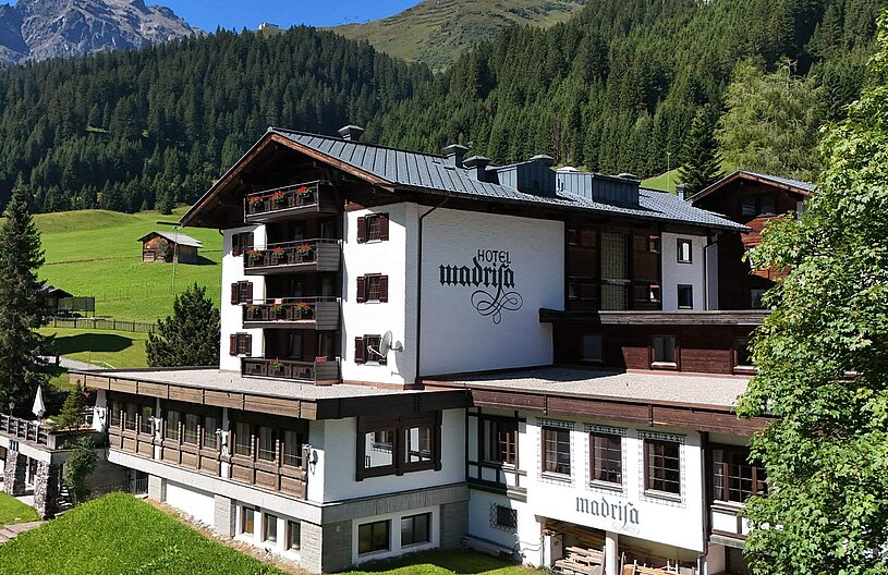 Das Bild zeigt das Hotel Madria, ein charmantes Gebäude mit mehreren Etagen und Balkonen, umgeben von üppigem Grün und Bergen im Hintergrund. Die Fassade ist in Weiß und Holz gehalten, mit dem Schriftzug „Hotel Madria“ deutlich sichtbar.