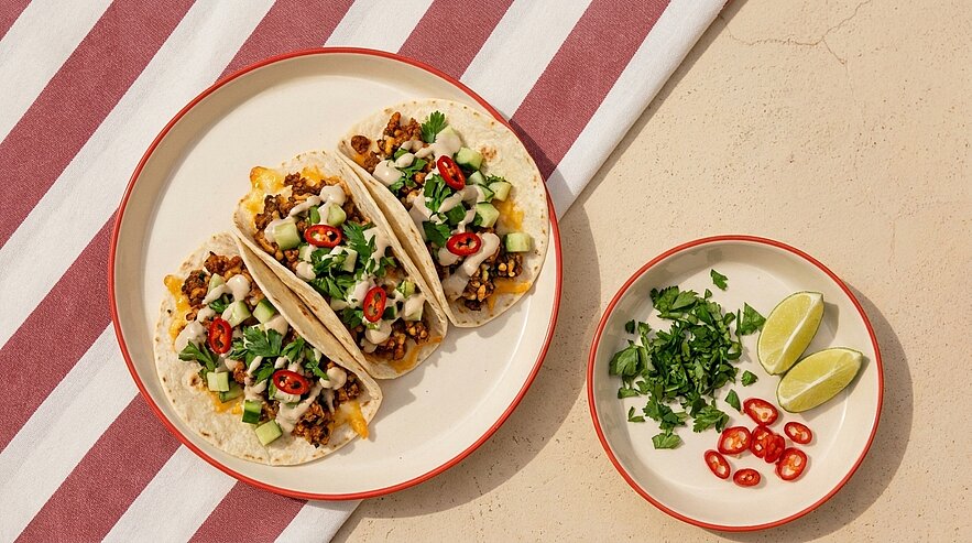 Drei Tacos mit Hackfleisch, geschmolzenem Käse, Avocadowürfeln, Koriander und roten Chili-Scheiben; daneben Schale mit Koriander, Limettenvierteln und Chili.