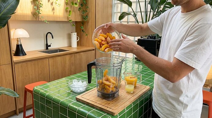 Hände einer Person schütten geschnittene Pfirsiche aus einer Schüssel in einen Standmixer auf einer Küchentheke; neben dem Mixer stehen ein Glas Saft und eine Schale Eiswürfel.
