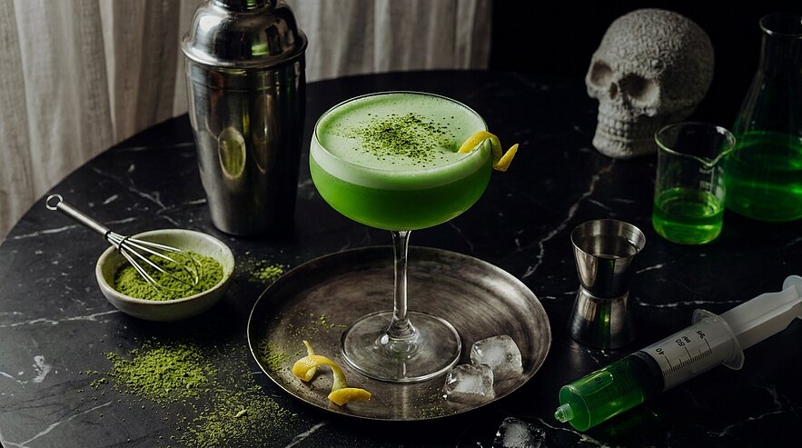 Grüner Matcha-Cocktail in einer Coupette, mit Zitronenzeste garniert, steht auf einem Metalltablett neben Eiswürfeln; Matchapulver ist verstreut, Barkeeper-Shaker und Messbecher im Hintergrund.