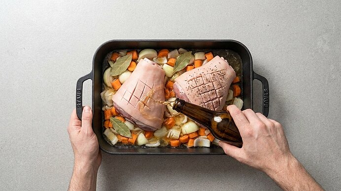 Zwei eingeschnittene Schweinebratenstücke in einer Auflaufform auf Karotten, Zwiebeln und Lorbeerblättern; Hände gießen Öl über das Fleisch.