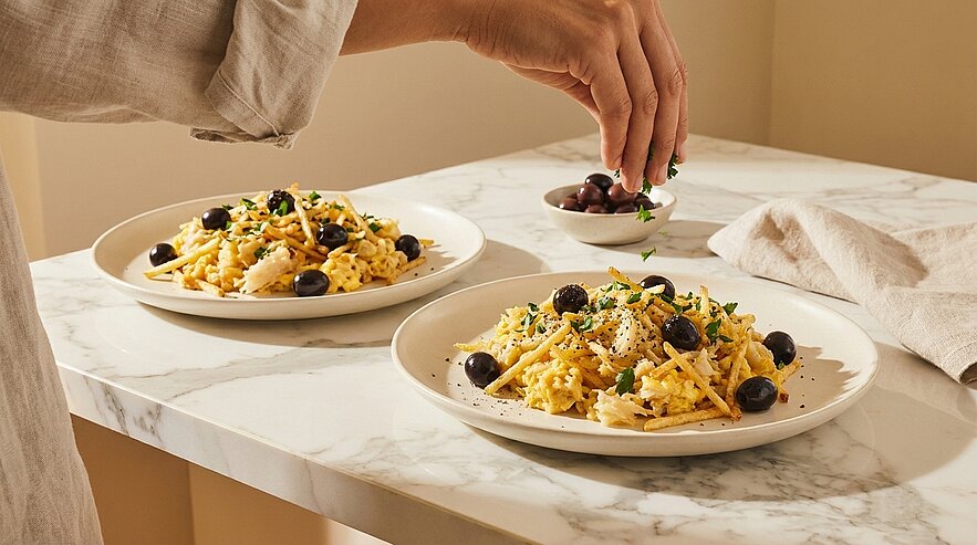 Zwei Teller Pasta mit schwarzen Oliven, geriebenem Käse und gehackten Kräutern auf einer Marmorarbeitsfläche; eine Hand streut Kräuter über einen Teller.