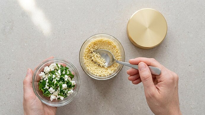 Zwei Hände bereiten Zutaten: links eine Schüssel mit gehackten Kräutern und zerbröseltem Käse, rechts eine Schüssel mit Couscous, in der ein Löffel rührt.