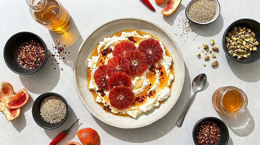 Schale mit cremigem Joghurt oder Quark, belegt mit leuchtend roten Blutorangenscheiben, Honig- oder Sirupdrizzle und Gewürzen; daneben Schälchen mit Pistazien, Sesam und Chiliflocken.