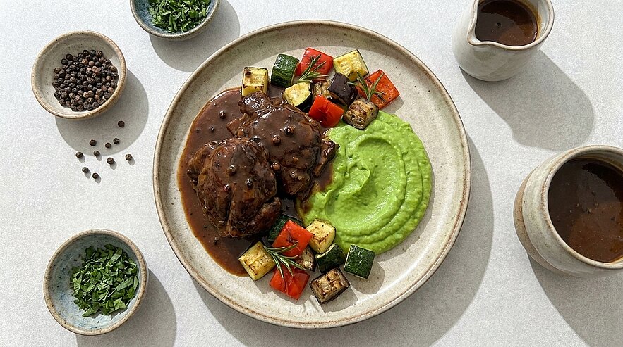 Teller mit zwei Fleischmedaillons in dunkler Pfefferrahmsauce, dazu grünes Erbsenpüree und buntes, gebratenes Gemüse (Zucchini, Paprika, Aubergine).