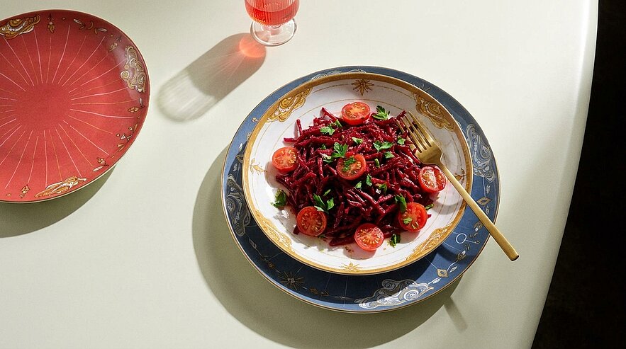 Vogelperspektive auf einen Teller rote Pasta mit halbierten Kirschtomaten und gehackten Kräutern, serviert auf dekorativen Tellern mit einer goldenen Gabel.