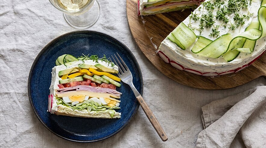 Anschnitt einer herzhaften, mehrlagigen Sandwich-Torte mit bunten Schichten aus Wurst, Käse und Gemüse als Stück auf Teller; Rest der dekorierten Torte mit Gurkenbändern und frischen Kräutern auf einem Holzbrett.