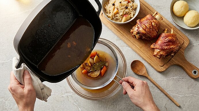 Hände sieben eine dunkle Brühe aus einem Bräter durch ein Sieb in eine Schüssel; daneben ein Holzbrett mit zwei knusprigen Schweinshaxen, eine Schüssel Sauerkraut und zwei Kartoffelknödel.