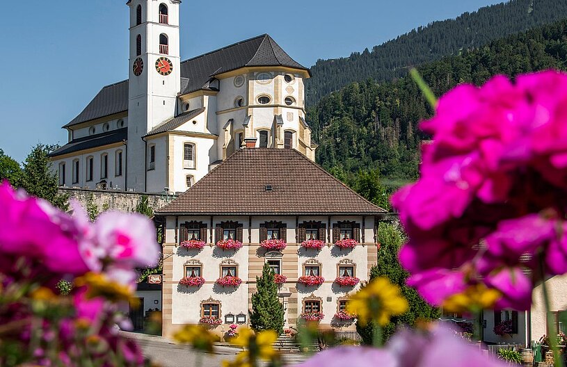 Eine malerische Ansicht eines traditionellen Gebäudes mit bunten Blumenkästen. Im Hintergrund erhebt sich ein Kirchturm. Die Umgebung ist von grünen Bäumen und Bergen umgeben, unter einem klaren blauen Himmel.