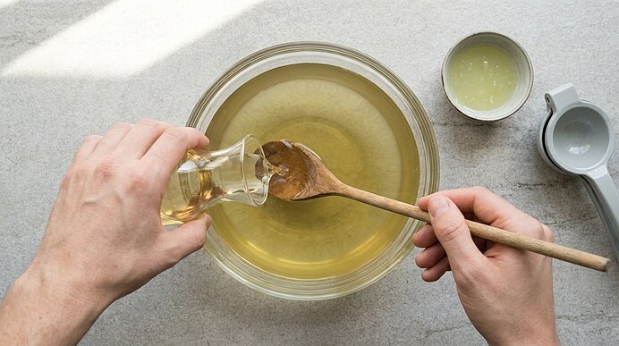 Zwei Hände gießen Holunderblütensirup über einen Holzkochlöffel in eine Glasschüssel mit grünem Tee.
