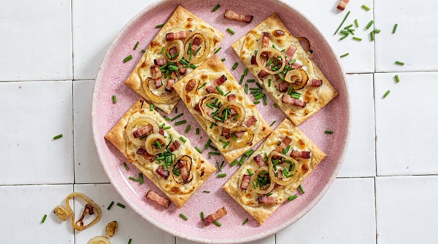 Mehrere rechteckige Blätterteig-Tartelettes auf einem rosa Teller, belegt mit karamellisierten Zwiebeln, knusprigem Speck und gehacktem Schnittlauch.