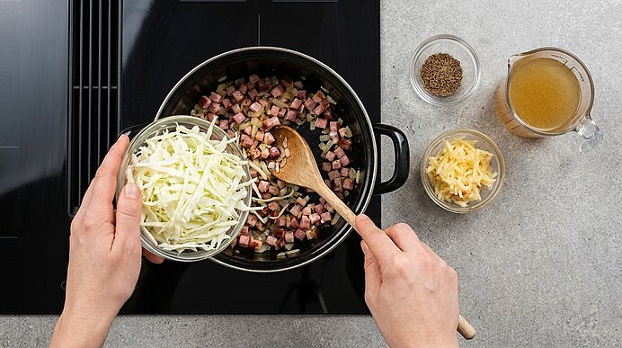 Hände geben gehobelten Weißkohl in einen Topf mit angebratenen Speckwürfeln, ein Holzlöffel rührt; daneben Schälchen mit Brühe, geriebenem Käse und Gewürzen.