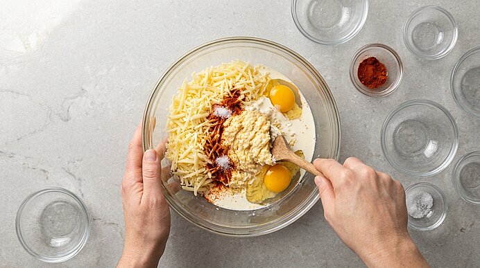Zwei Hände verrühren geriebenen Käse, Eigelb, Sahne, Paprikapulver und Salz mit einem Holzlöffel in einer Glasschüssel.