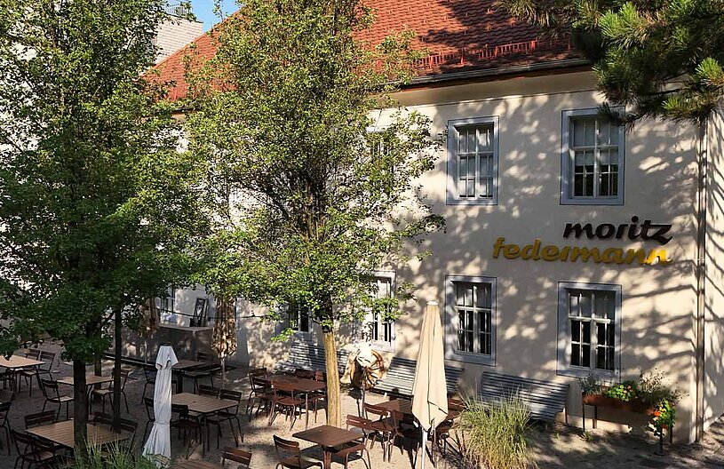 Das Bild zeigt ein Restaurant mit Außenbereich, umgeben von Bäumen. Mehrere Tische und Stühle sind im Freien aufgestellt, einige Sonnenschirme sind geschlossen. An der Wand des Restaurants steht der Name "Moritz Federmann" in Verbindung mit hellem Licht.