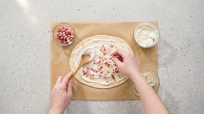 Zwei Hände belegen einen mit Creme bestrichenen Flammkuchen auf Backpapier mit Zwiebelringen und Speckwürfeln.