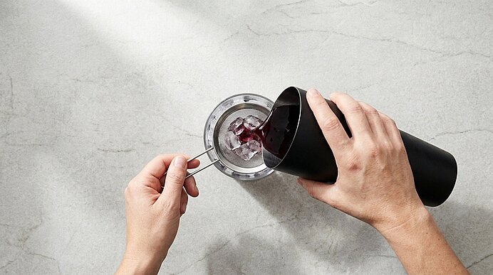 Draufsicht: Zwei Hände sieben dunkelrote Flüssigkeit aus einem schwarzen Cocktail-Shaker durch ein feines Sieb in ein Metall-Cocktailglas.