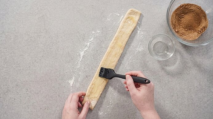Zwei Hände bestreichen einen schmalen Teigstreifen mit Margarine. Daneben steht eine Schüssel mit Zimt-Zucker.